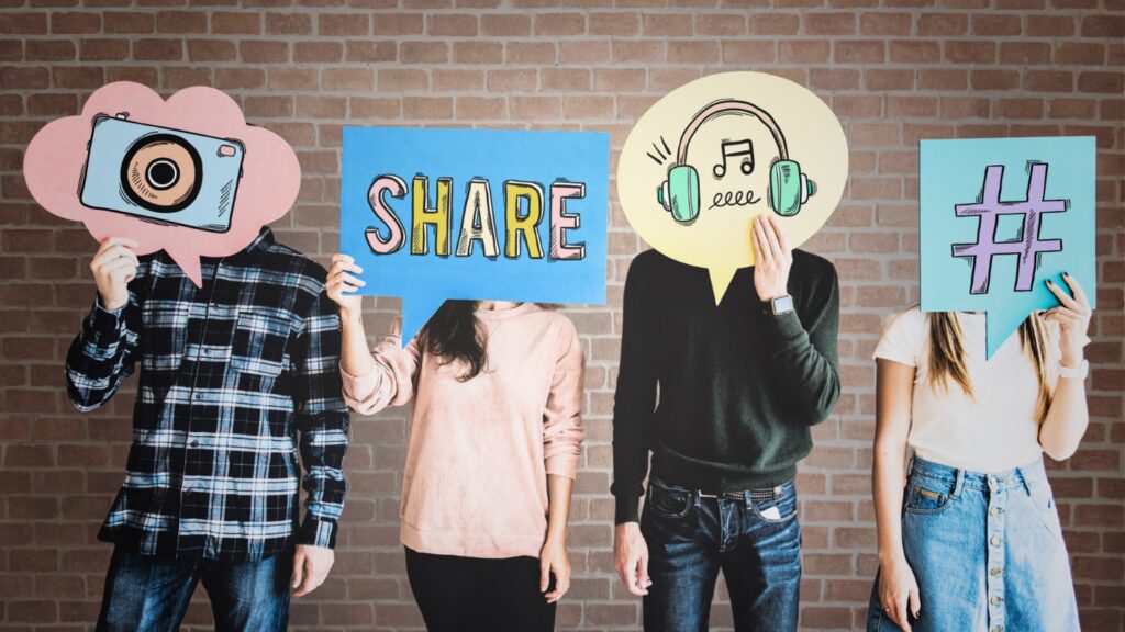 This picture shows four people standing side by side in front of a brick wall. Each person is holding a large, colorful sign in front of their face, so their faces are hidden. The signs are shaped like speech bubbles and contain symbols or words that represent social media and communication.
